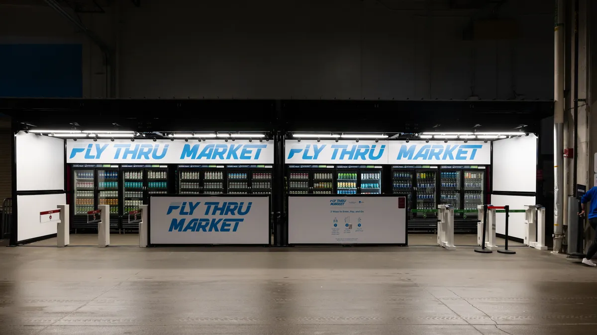 A photo of a concession stand in a sports arena. It includes bankds of coolers and several gates for entry and exit. Signs on a call in front and the wall above the coolers all say "Fly Thru Market."