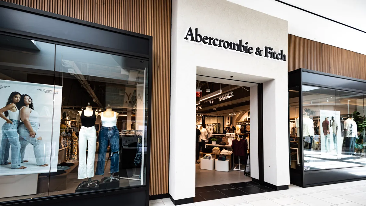 Storefront of Abercrombie & Fitch in a mall. Store windows on the left and right have mannequins wearing long pants and shirts.