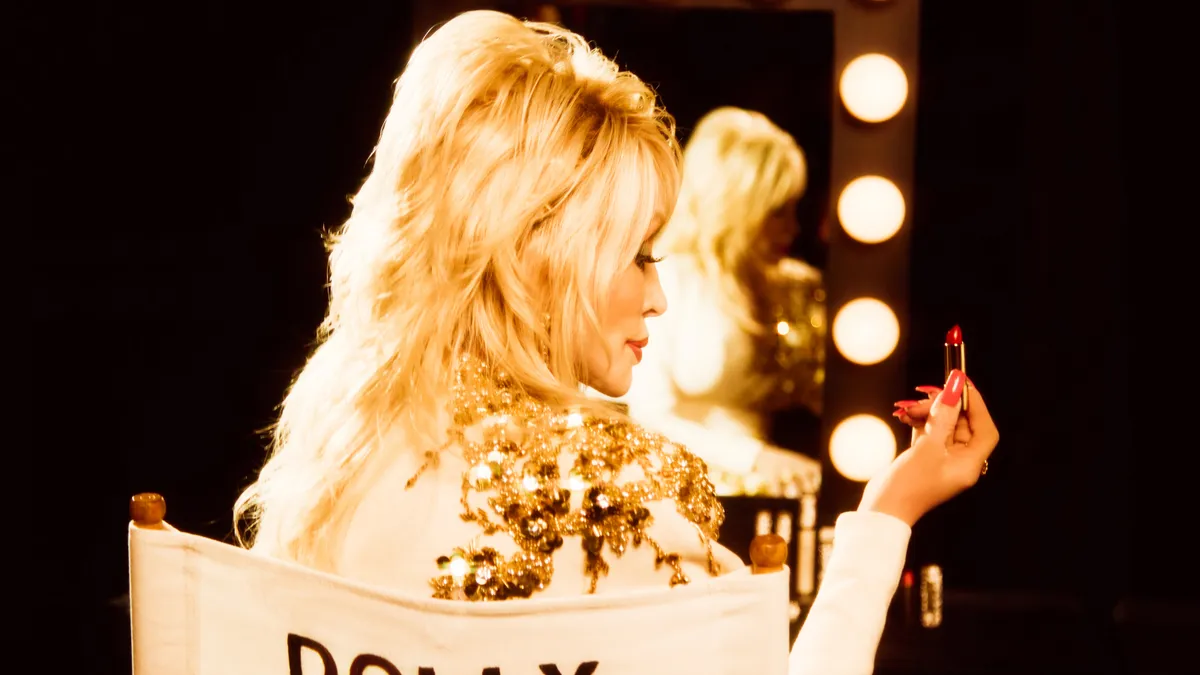 A person, Dolly Parton, is seen from behind holding a lipstick and sitting in chair that reads "Dolly" across the back.