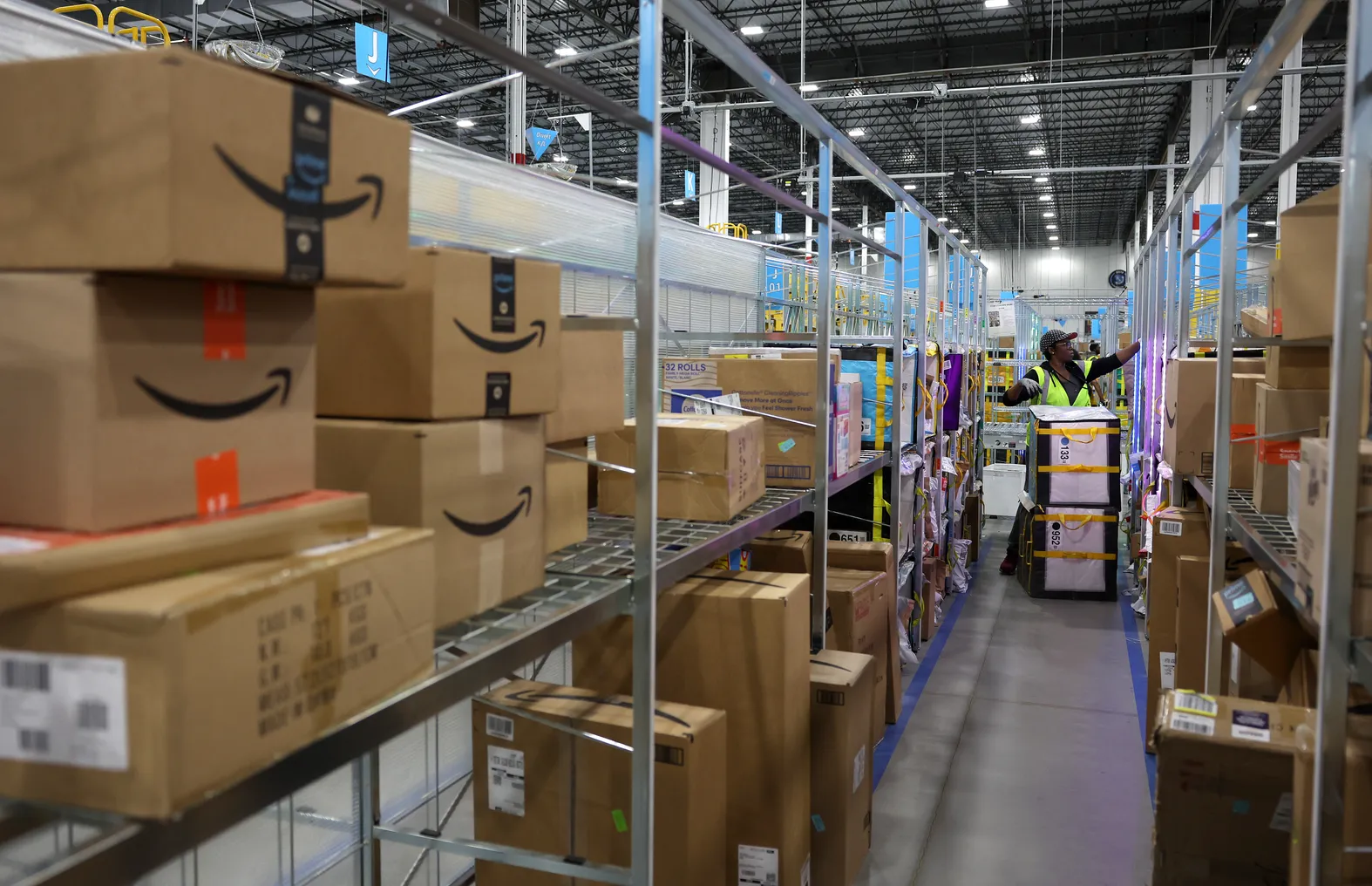 An Amazon worker with a cart sorts packages in a warehouse aisle packed with Amazon packages.