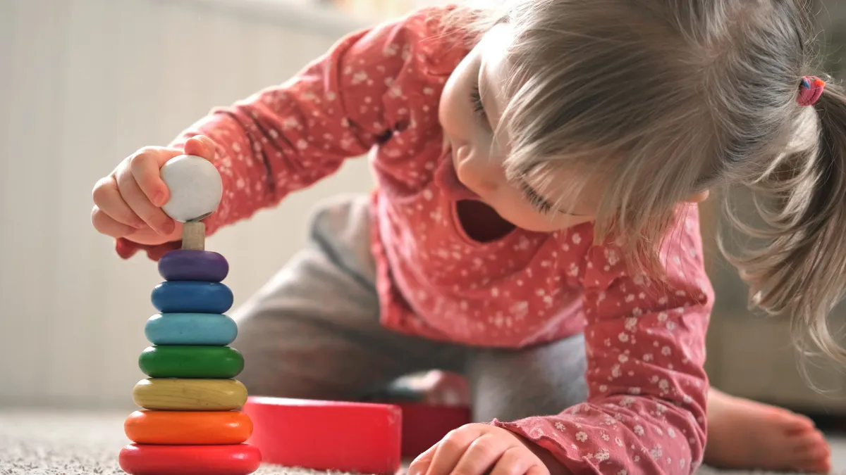 A young students in on the floor with head bent down toward and touching a colorful wood ring toy.