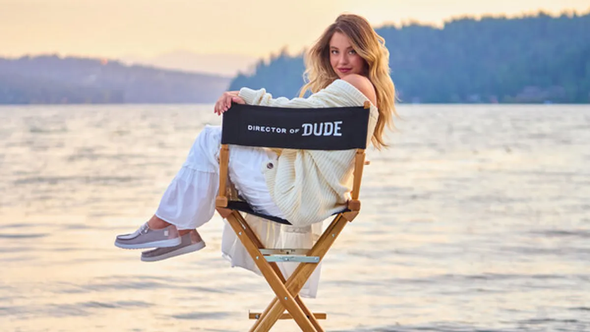 A person with long blond hair smiles, sitting sideways in a director-style chair. Water and far-off trees are in the background.