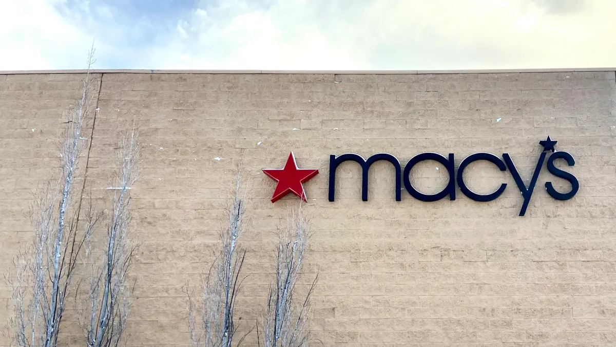 Four birch trees to left of a light brick wall with a red star and a "Macy's" sign, with a small row of bushes in snow.